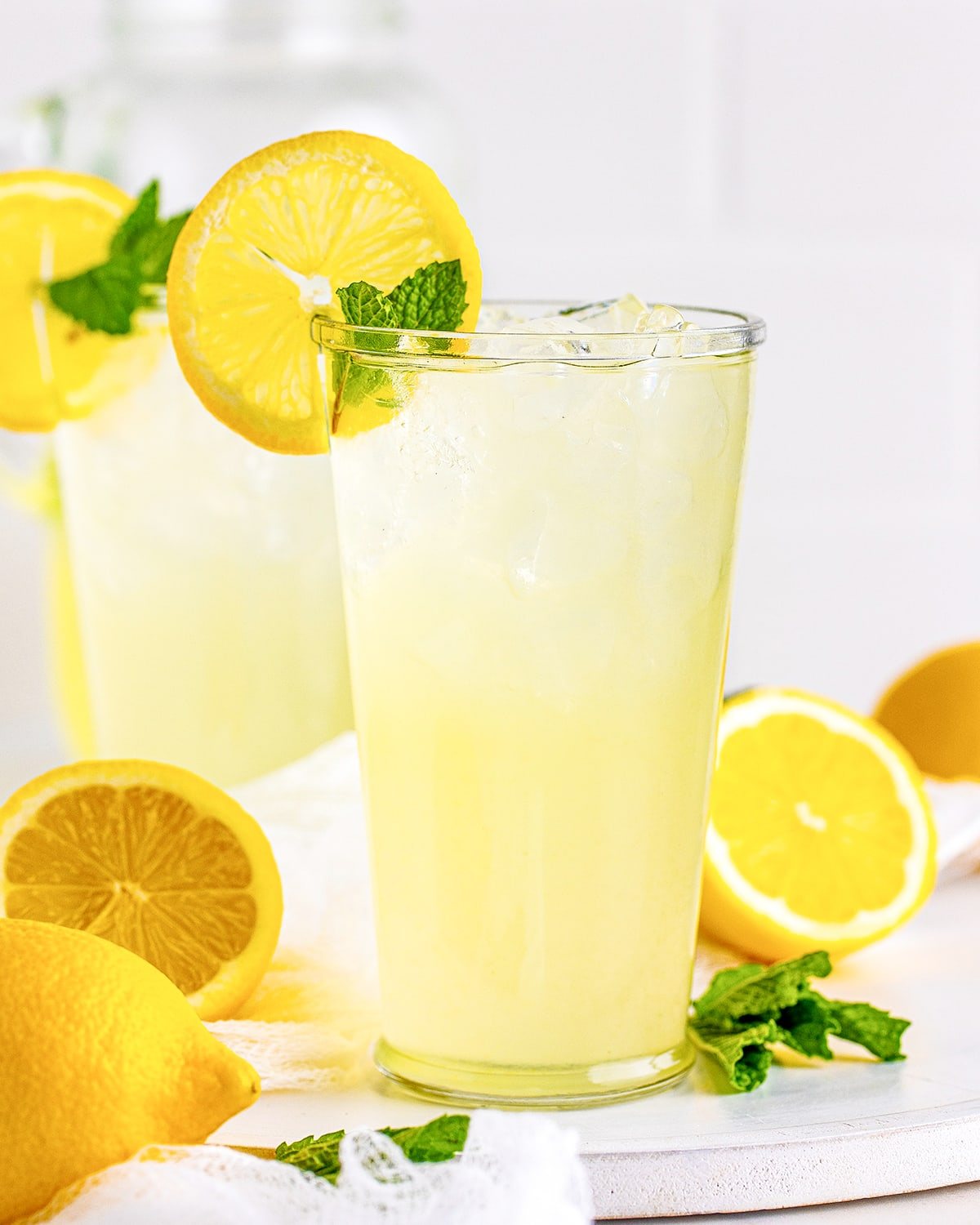 A tall glass of fresh-squeezed lemonade with ice, a lemon slice and mint, with halved lemons around it